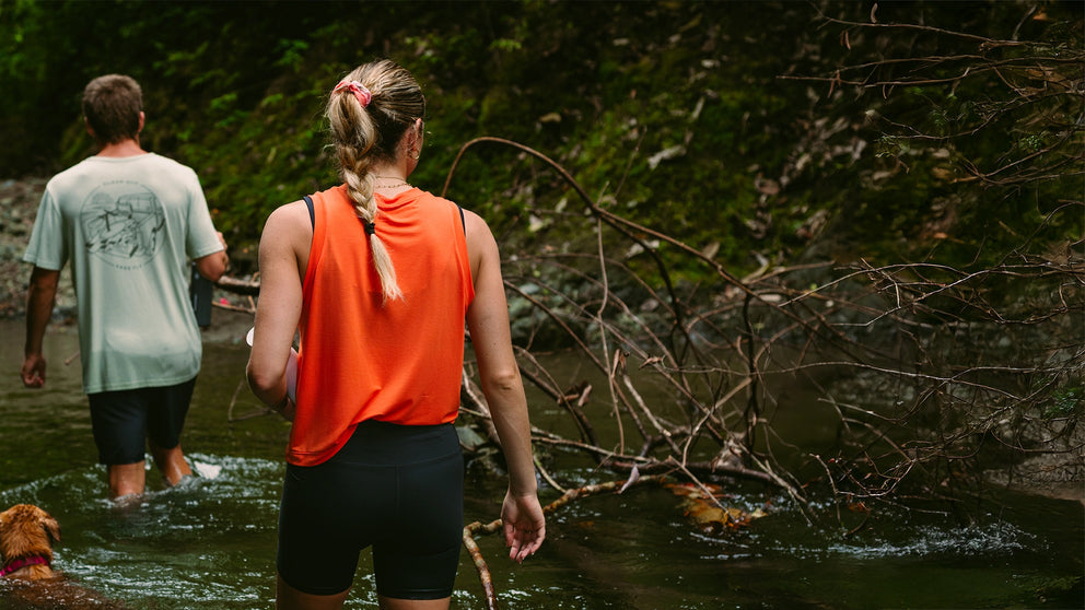 Two people and a dog wading through a stream in a natural setting